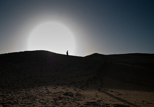 Fotografia de uma duna com o sol em fundo e uma pessoa no topo da duna