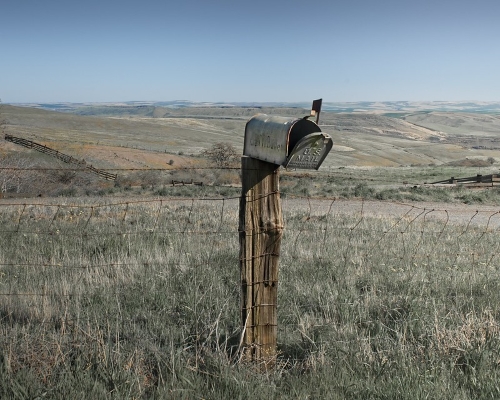 Fotografia de um correio abandonado no meio do campo circundado por uma rede de ferro